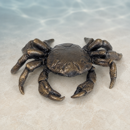 A realistic metal crab figurine with a textured bronze finish, positioned on a light beige stone surface with a soft, blurred water-like blue background.