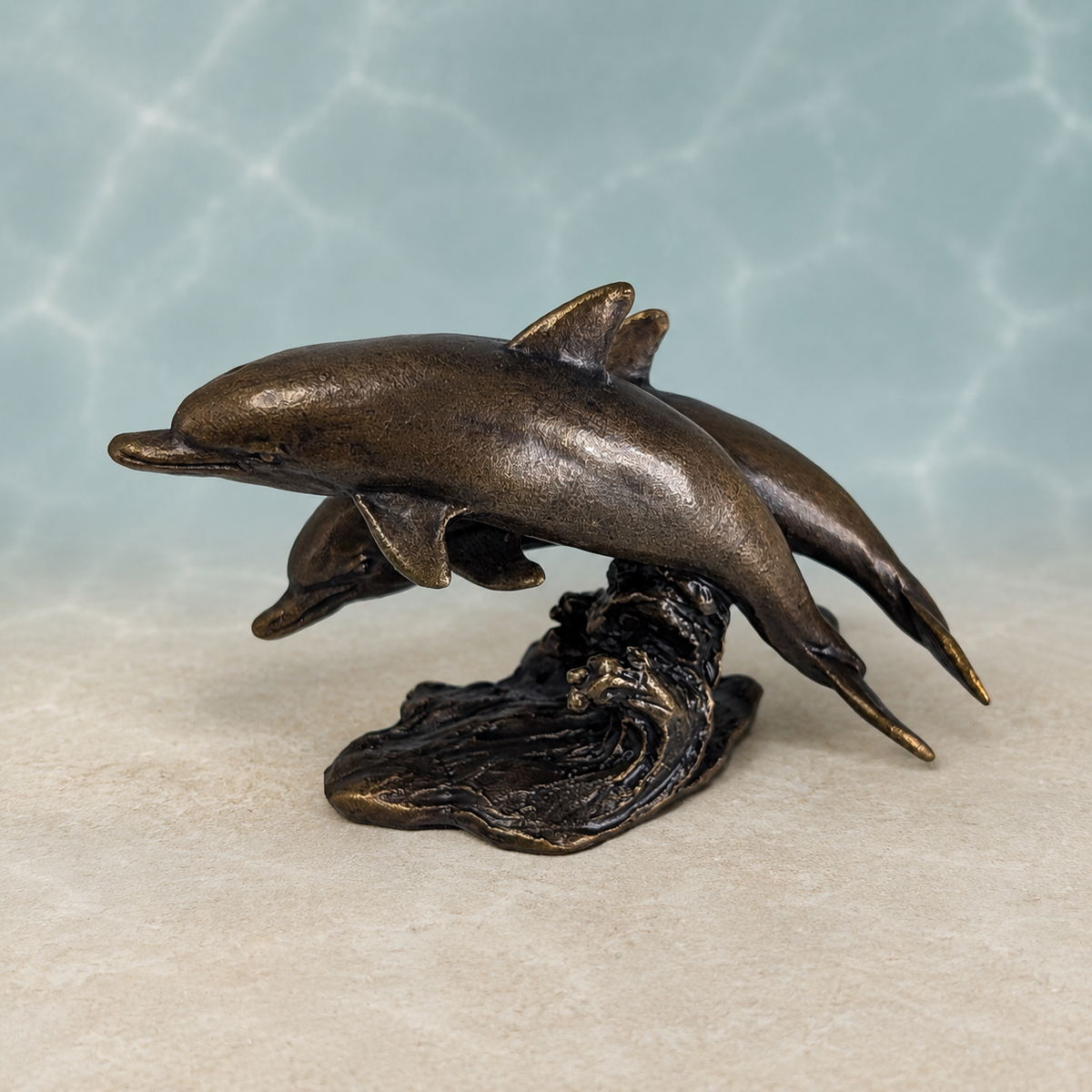 Bronze-finished sculpture of two dolphins leaping from above a wave base, displayed against a light blue water-ripple backdrop and beige surface.