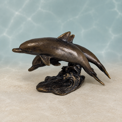 Bronze-finished sculpture of two dolphins leaping from above a wave base, displayed against a light blue water-ripple backdrop and beige surface.