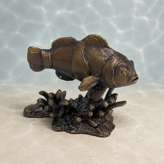 Bronze-finished fish sculpture standing on a coral-like base, photographed against a light blue water-textured backdrop on a pale surface.