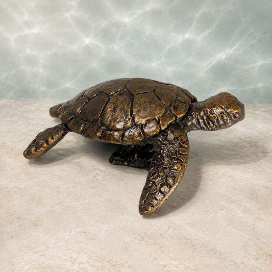 Bronze sea turtle figurine with textured shell and flippers, displayed on a light beige stone surface against a water-ripple-patterned background.