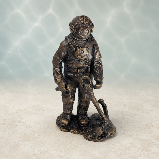 A small bronze-colored figurine of a vintage diving suit with a helmet and scuba gear, standing on a textured base against a light blue-green water ripple background.