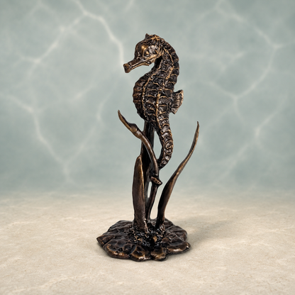 A bronze-finished seahorse sculpture standing on a stylized underwater rock and seaweed base, photographed against a soft blue-green water-like backdrop with light reflections.