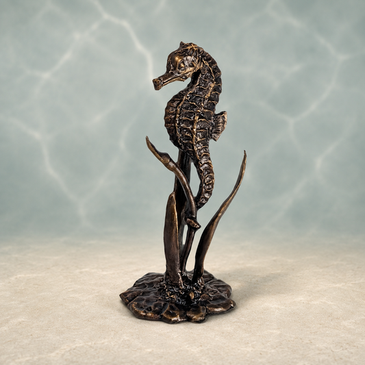 A bronze-finished seahorse sculpture standing on a stylized underwater rock and seaweed base, photographed against a soft blue-green water-like backdrop with light reflections.