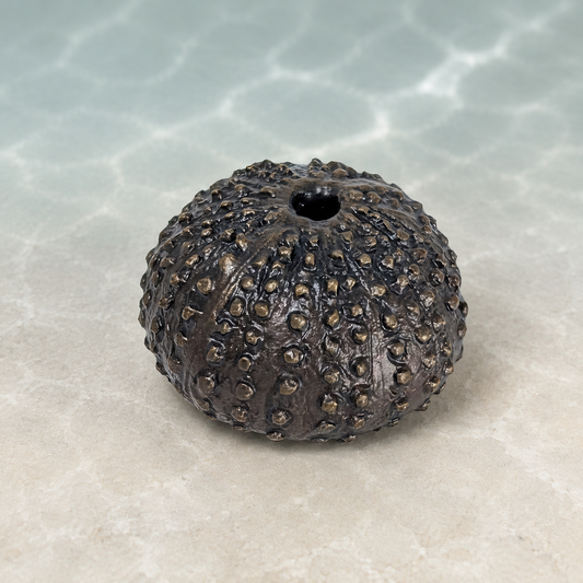 A small spherical, dark metallic vessel with a heavily textured, bumpy surface and a central hole, resembling a miniature sea urchin or bead, shown on a light beige stone surface with a blue-green water-like background.