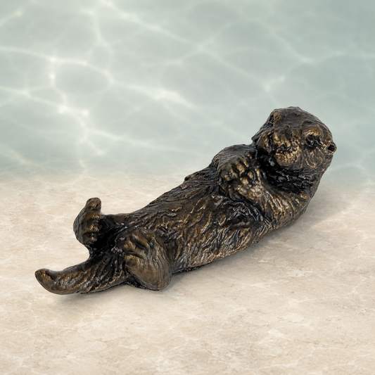 A bronze-finished sculpture of a sea otter lying on its back, set against a background with a light beige floor and a soft, water-like blue and white textured backdrop.