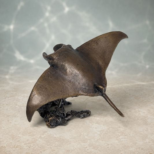A bronze sculpture of a manta ray resting on a dark rocky base, displayed on a textured beige surface with a soft, water-like blue background.
