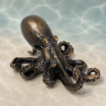 A small bronze octopus figurine with textured, metallic surfaces, displayed on a light beige surface with a soft, water-like blue background.