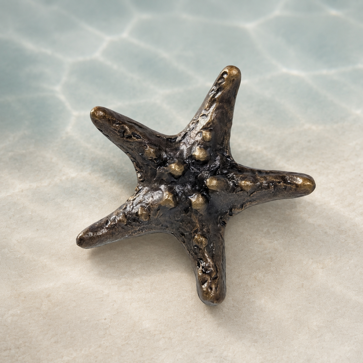 A small five-pointed starfish figurine with a textured, metallic bronze finish, resting on a light beige surface with a soft-focus background resembling shallow water.
