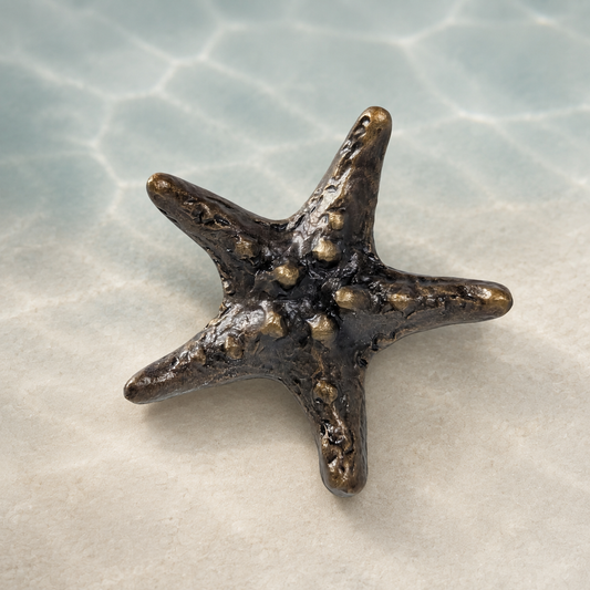 A small five-pointed starfish figurine with a textured, metallic bronze finish, resting on a light beige surface with a soft-focus background resembling shallow water.