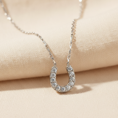 A delicate silver chain necklace featuring a horseshoe-shaped pendant paved with small clear stones, set against a plain white background.