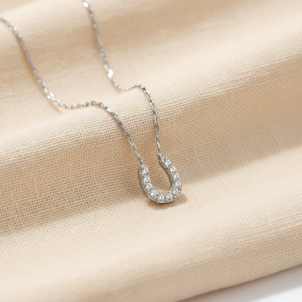 A delicate silver chain necklace featuring a horseshoe-shaped pendant paved with small clear stones, set against a plain white background.