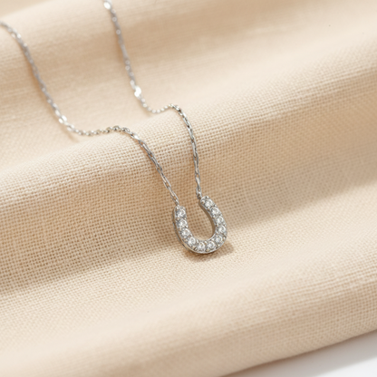 A delicate silver chain necklace featuring a horseshoe-shaped pendant paved with small clear stones, set against a plain white background.