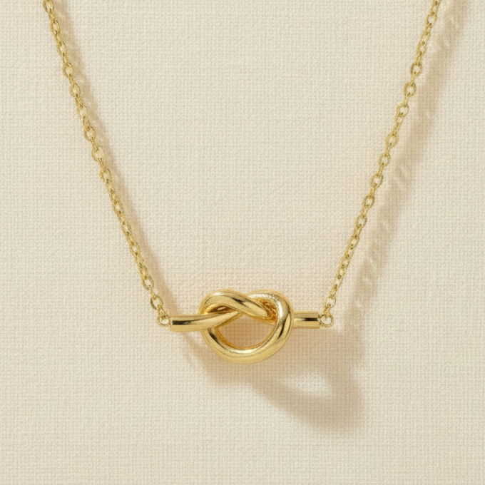 A gold necklace featuring a polished knot pendant with a flat bar extending from each side, suspended from a delicate link chain against a cream cotton background.