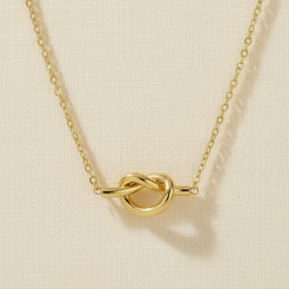 A gold necklace featuring a polished knot pendant with a flat bar extending from each side, suspended from a delicate link chain against a cream cotton background.